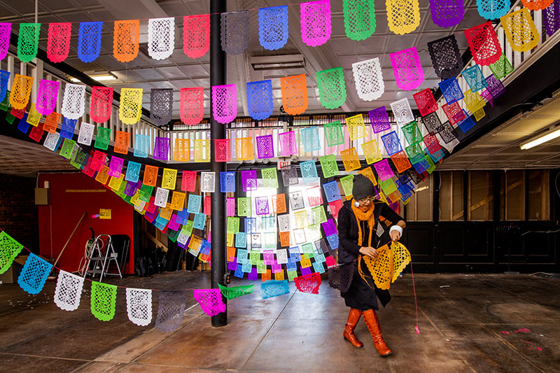 Blanka Amezkua installing colorful papel picado designs near ceiling of large space. Photo by Meredith Mashburn