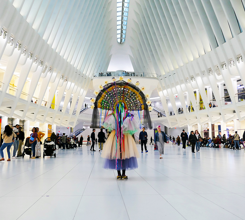 Blanka Amezkua in contemporary Mexican indigenous and plant inspired costume; wearing large headress, view from back. She is standing amidst a crowd filled imposing white Oculus station in NYC. Performance piece for Art in Odd Places festival. Photo by Leo A